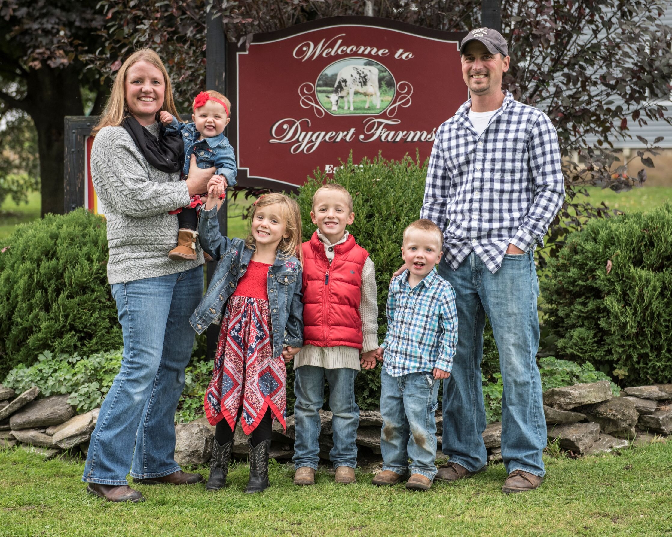 Family posing in front of the Dygert Farms welcome sign, featuring a mother holding a baby, three children in colorful outfits, and a father in a plaid shirt, all set against a lush green background.