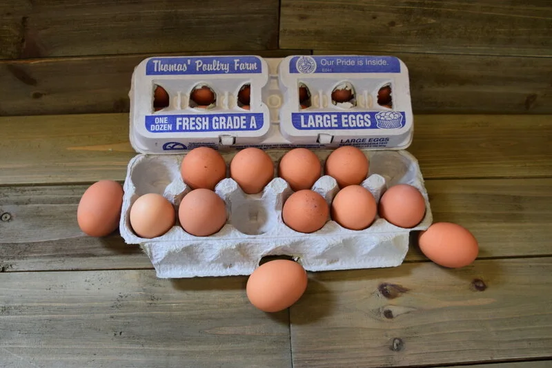Fresh Grade A large eggs from Thomas' Poultry Farm displayed in an egg carton on a wooden surface, showcasing a dozen eggs with a few outside the carton.
