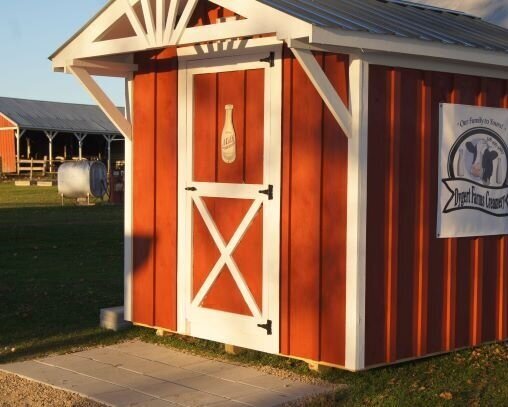 Red barn-style shed with white trim and a decorative door, featuring a milk bottle illustration, set in a rural landscape with additional outbuildings in the background.